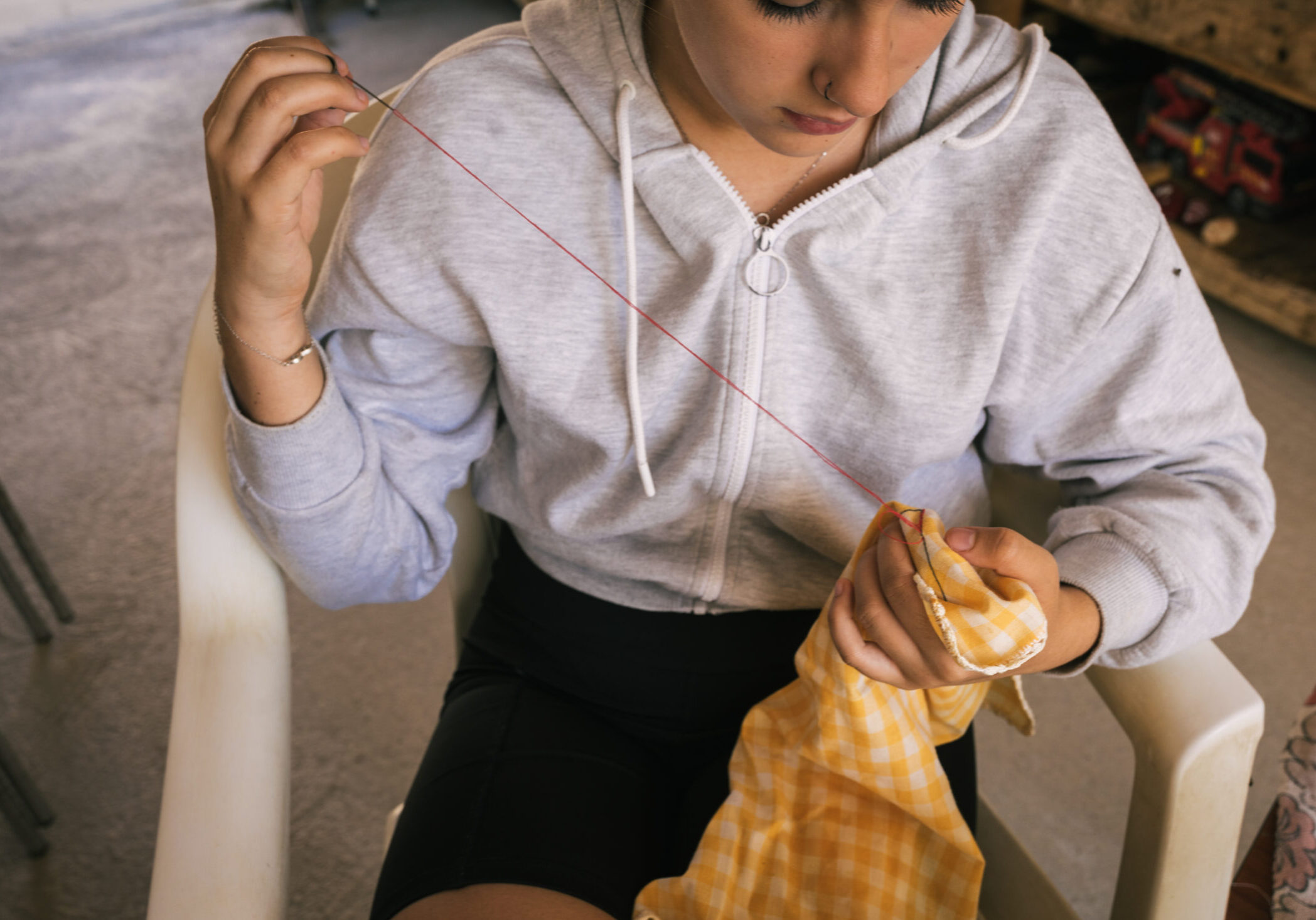 brunette teenage girl in gray sweatshirt is sewing a red heart with thread on a yellow scarf